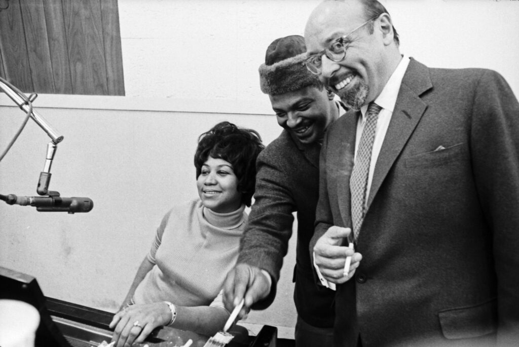 Black and white photo showing Aretha Franklin, Ted White and Ahmet Ertegun in Atlantic Studios. Aretha is seated in front of a microphone, her husband Ted White is standing next to her and leaning forward, and Ahmet Ertegun is standing on the right of the image looking at the camera and holding a cigarette in his right hand